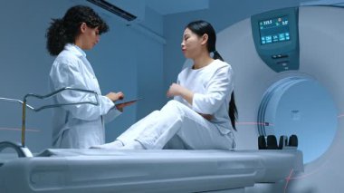 Modern ekipmanların arka planındaki manyetik rezonans görüntülerinin incelenmesi. Bilgisayarlı tomografi tarama yatağında oturan hasta tüm vücut ağrısından şikayetçi, dikkatle doktoru dinliyor..