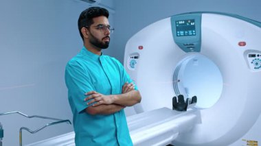 Hintli tomografi uzmanı çapraz kollarla poz veriyor. Tomografinin arka planında ciddi bir doktor var. Müslüman doktor gözlük takıyor. MRI incelemesi için modern donanımlı oda. MRI kapsülü.