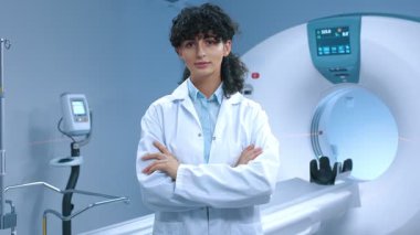 Genç tomografi uzmanı kollarını kavuşturmuş poz veriyor. Tomografi tarayıcısının arka planında dost bir doktor var. MRI incelemesi için modern donanımlı oda. MRI kapsülü. MRI uzmanı. Sağlık çalışanı.