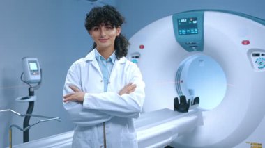 Genç tomografi uzmanı kollarını kavuşturmuş poz veriyor. MRI kapsülünün arka planında gülümseyen neşeli doktor. MRI incelemesi için modern donanımlı oda. MRI uzmanı. Sağlık çalışanı.