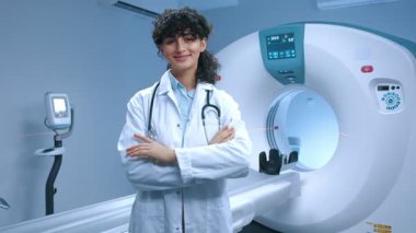 Genç tomografi uzmanı kollarını kavuşturmuş poz veriyor. MRI cihazlarının arka planında gülümseyen neşeli bir doktor. MRI incelemesi için kabine. MRI uzmanı. Steteskoplu bir tıp çalışanı..