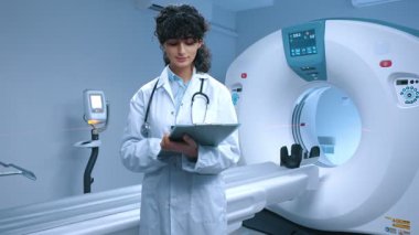 Genç tomografi uzmanı ameliyata hazırlanıyor. MRI cihazlarının arka planında gülümseyen neşeli bir doktor. MRI incelemesi için kabine. MRI uzmanı. Steteskoplu bir tıp çalışanı..