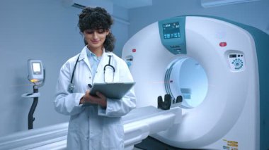 Genç tomografi uzmanı ameliyata hazırlanıyor. MRI cihazlarının arka planında gülümseyen neşeli bir doktor. MRI incelemesi için kabine. MRI uzmanı klasörü tutuyor. Steteskoplu bir tıp çalışanı..