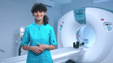 Genç tomografi uzmanı geniş bir gülümsemeyle poz veriyor. MRI cihazlarının arka planında gülümseyen neşeli bir doktor. MRI incelemesi için kabine. MRI uzmanı. Tomografi teknisyeni. Tıbbi uzman..