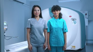 Mavi üniformalı nörobilimci doktorlar işyerindeki kameraya bakıyor. MRI kapsülünün arka planında nöro-adiyolojik işçiler, tıbbi x-ray makinesi kullanıyorlar..