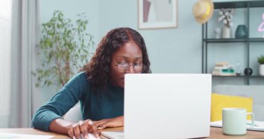 Masada oturmuş gülümserken dizüstü bilgisayara bakan neşeli Afro-Amerikalı güzel kız. Bilgisayarda okuyan tatmin olmuş duygusal kız öğrencinin portresi. Mesafe çalışma kavramı