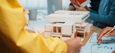 Bir grup profesyonel, tasarım planları ve modern ofis ortamında ayrıntılı bir model üzerinde çalışan bir mimari proje üzerinde işbirliği yapmaktadır. SACTR