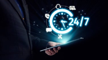 Profesyonel işadamı, dijital çağda 24 saat kullanılabilirliği ve iletişimi temsil eden ve sürekli desteği sembolize eden parıldayan bir saat simgesine sahip bir tablet tutuyor. Dörtgen