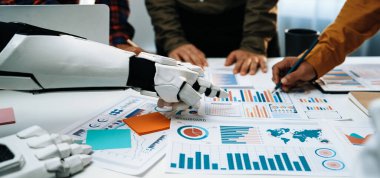 Robotik bir el, verileri analiz eden ve grafikler ve grafiklerle stratejiler geliştiren, modern ofislerde teknoloji ve takım çalışmasının karışımını gösteren bir iş ekibine yardım eder. Noogenesis