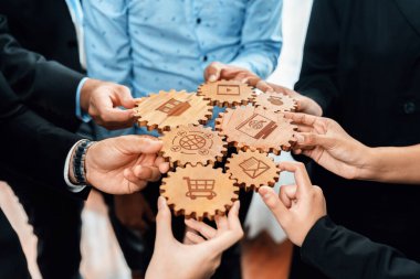 A diverse group of professionals holds wooden gears, each engraved with business-related icons. This image symbolizes collaboration, strategy, and innovation in a modern workplace setting. Amity