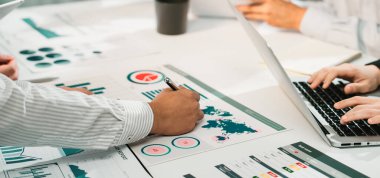 Profesyonel bir ekip, bir iş toplantısı sırasında, teknoloji ve belgeler masaya saçılmış vaziyette mali raporları ve veri tablolarını analiz ederek işbirliği yapıyor. SACTR