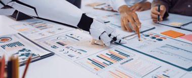 Grafikler ve grafikler içeren çeşitli veri analiz belgeleri üzerinde çalışan bir insan ve robotu gösteren modern bir ofis sahnesinin yakından bir görüntüsü.