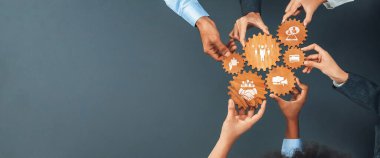 A group of diverse individuals gathers around to hold wooden gears with icons, symbolizing teamwork and business strategies, portraying collaboration in the modern workplace. Amity
