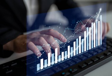 A focused business professional interacts with a keyboard while viewing financial graphs and data trends on a transparent display in a modern office setting. Impute