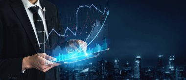 Resmi takım elbiseli bir işadamı, dijital grafik ve grafiklerle çevrili mali verileri analiz etmek için bir tablet kullanır ve gece arkaplanda kentsel bir ufuk çizgisi bulunur. Kafa derisi