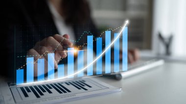 Bir işletme profesyoneli masadaki finansal tablolar ve grafiklerle etkileşime girer, büyüme eğilimlerini gösterir ve modern bir çalışma alanında stratejik planlamayı vurgular. Etki
