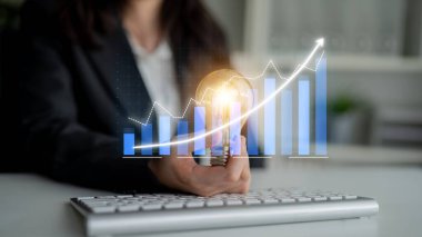 Profesyonel bir kadın modern bir ofisteki veri eğilimlerini analiz ederken, iş ortamındaki yenilik ve büyümeyi sembolize eden bir ampul tutuyor. Etki