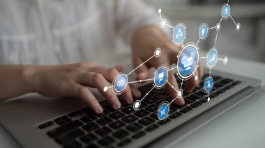 Modern bir çalışma alanında çeşitli teknoloji ve iletişim temalarını sembolize eden parıldayan dijital simgelerle çevrili bir dizüstü bilgisayarda yazı yazan ellerin yakın görüntüsü. Tessel