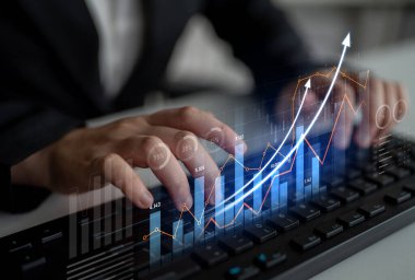 Bir iş adamı klavyede yazarken finansal verileri analiz eder. Grafikler ve grafikler büyüme eğilimlerini ve performans ölçümlerini gösteriyor, pazar anlayışlarını vurguluyor. Kafa derisi
