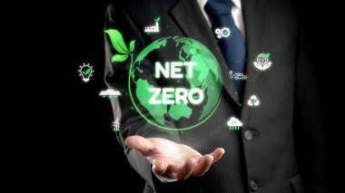 Bir işadamı, modern uygulamalar ve girişimlerdeki sürdürülebilirlik ve eko-arkadaşlığı gösteren yeşil simgelerle çevrili net sıfır hedefleri sembolize eden bir dünya sunuyor. Dörtgen
