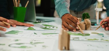 Dinamik bir çalışma alanında, sürdürülebilir tasarım kavramlarını analiz eden ve çizen, takım çalışmalarını ve çevre dostu stratejileri vurgulayan çeşitli bireylerin elleri sergileniyor. SACTR