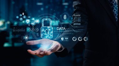 Bir iş adamı elinin üstünde çeşitli teknolojik unsurlarla dijital veri güvenliği ikonu sunarak modern ofis ortamında korunmayı ve yeniliği sembolize ediyor. Dörtgen