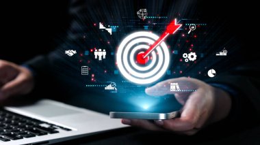 Bir iş adamı akıllı bir telefonla iletişim kurar, dijital ikonlar aracılığıyla hedefleri görselleştirir ve dinamik bir iş ortamında strateji, başarı ve teknolojiyi temsil eder. Dörtgen