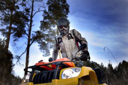 Photo for Horizontal close-up of a man in helmet and safety goggles looking into the camera while sitting on quad bike against vivid blue sky. - Royalty Free Image