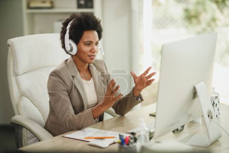 Foto de Fotografía de una atractiva empresaria africana con auriculares que recibe videollamadas mientras trabaja en la computadora durante la pandemia del virus corona. - Imagen libre de derechos