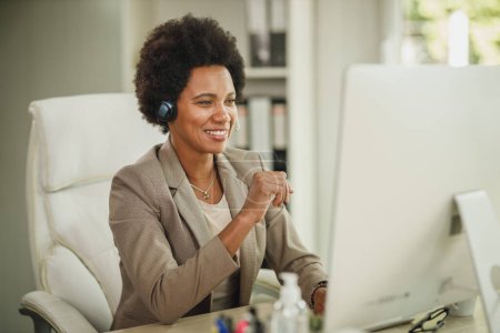 Aufnahme einer attraktiven afrikanischen Geschäftsfrau, die während der Coronavirus-Pandemie allein in ihrem Büro mit Kopfhörern sitzt und am Computer arbeitet.