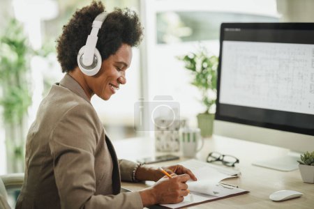 Photo for Shot of an African businesswoman wearing a headphones while working on her computer during corona virus pandemic. - Royalty Free Image