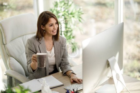 Una atractiva mujer de negocios bebiendo café mientras usa una computadora en su oficina en casa.