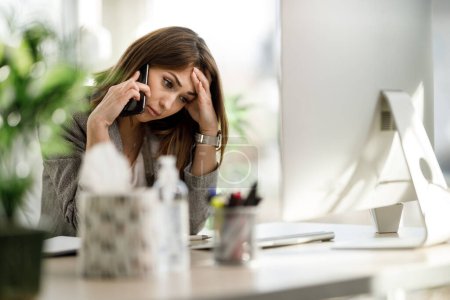 Eine müde Geschäftsfrau, die allein in ihrem Home Office sitzt und besorgt auf dem Smartphone spricht.