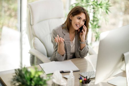 Eine lächelnde Geschäftsfrau spricht am Smartphone, während sie in ihrem Home Office am Computer arbeitet.