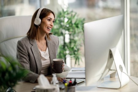 Eine lächelnde Geschäftsfrau genießt eine Tasse Kaffee, während sie im Home Office am Computer arbeitet.