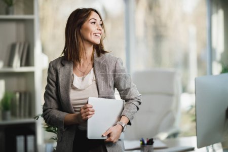 Une jeune femme d'affaires réussie tenant un ordinateur portable et debout dans son bureau.