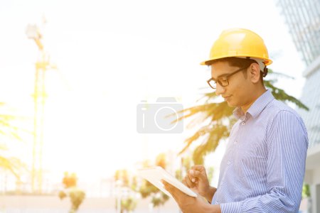 Foto de Ingeniero contratista masculino indio asiático con la mano del sombrero duro que sostiene la computadora de la tableta que inspecciona en el sitio de construcción, luz del sol de la mañana en el fondo
. - Imagen libre de derechos