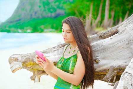 Téléchargez les photos : Belle adolescente utilisant un téléphone portable sur la plage hawaïenne, debout à côté du bois flotté - en image libre de droit