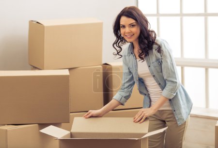 Photo for Attractive young woman is moving, standing among cardboard boxes, looking at camera and smiling - Royalty Free Image