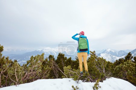 Foto de Vista posterior de la joven mujer con sombrero rosa, chaqueta azul, mochila verde, pantalones amarillos y botas de senderismo de pie contra el valle de la montaña de invierno y mirando lejos cubriendo sus ojos a mano - concepto de aventura - Imagen libre de derechos