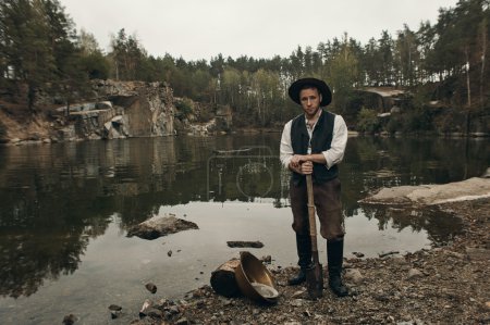 Photo for Caucasian gold digger in retro clothes standing near lake after hard work. He wears shirt, leather pants and boots.  Sky is cloudy and grey. He is near gold mining equipment. - Royalty Free Image