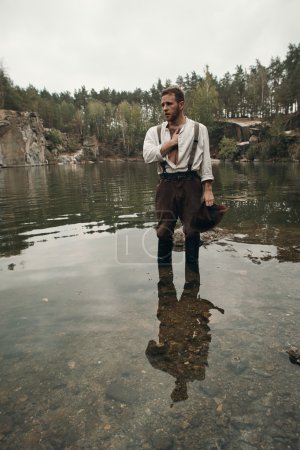 Téléchargez les photos : Creuseur d'or caucasien en vêtements rétro debout sur la roche dans le lac après un travail acharné. Il porte une chemise, un pantalon en cuir et des bottes. Ciel nuageux et gris
. - en image libre de droit