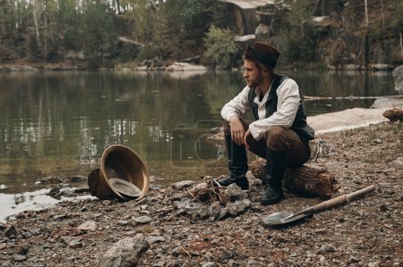 Photo for Caucasian gold digger in retro clothes warming up near fire after hard work. He wears shirt, leather pants, vest and boots.  Sky is cloudy and grey. There is gold mining equipment in the frame. - Royalty Free Image