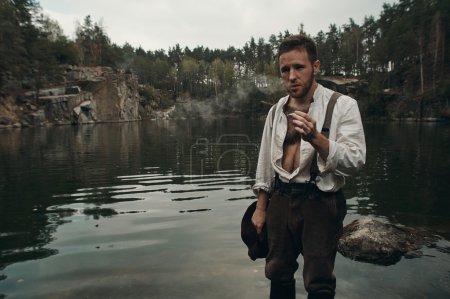 Téléchargez les photos : Creuseur d'or caucasien en vêtements rétro fume cigarette tout en se tenant dans le lac avec banque rocheuse après un travail acharné. Il porte une chemise, un pantalon en cuir, des galons et des bottes. Ciel nuageux et gris
. - en image libre de droit