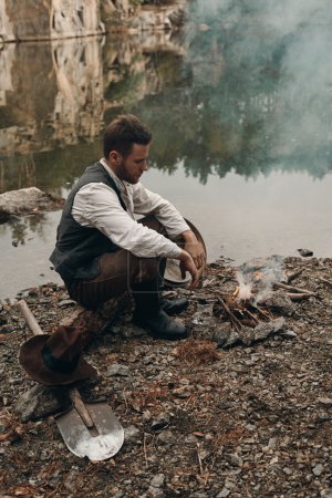 Foto de Excavadora de oro caucásico en ropa retro calentamiento cerca del fuego después del trabajo duro. Lleva camisa, pantalones de cuero, chaleco y botas. El cielo está nublado y gris. Hay equipos de minería de oro en el marco
. - Imagen libre de derechos