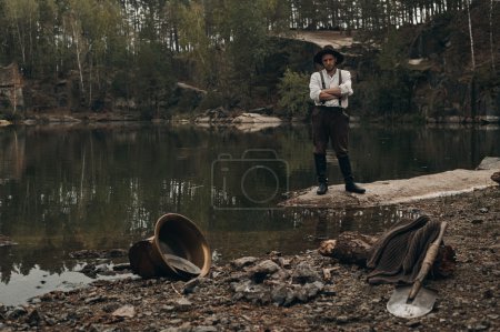 Photo for Caucasian gold digger in retro clothes standing near lake after hard work. He wears shirt, leather pants and boots.  Sky is cloudy and grey. He stands behind fireplace and gold mining equipment. - Royalty Free Image