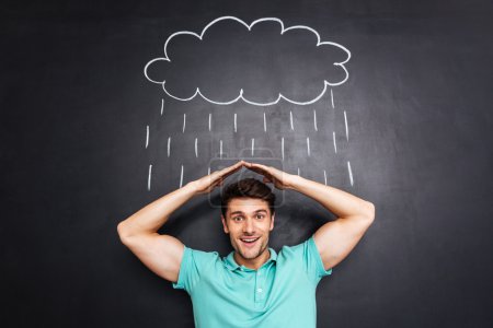 Photo for Smiling young man holding hands above head and covering from drawn rain over chalkboard background - Royalty Free Image