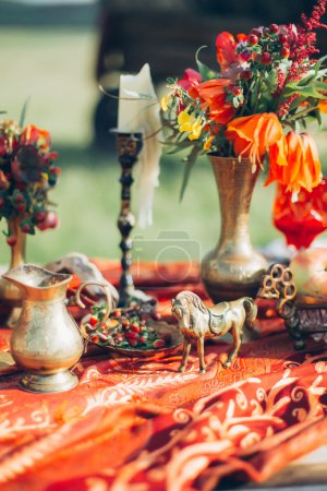 Téléchargez les photos : Table de salle à manger de style bohème, décorée avec des bougies, des fleurs et des articles en bronze. Photo prise dans le style des beaux-arts. Film stylisé. Belle table boho à l'extérieur sur un fond d'herbe verte
. - en image libre de droit