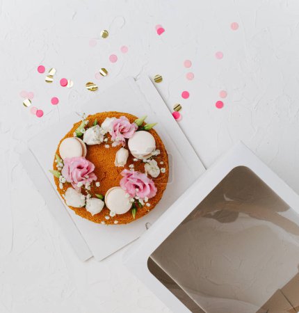 Vista superior de un pastel de miel con flores y macarrones en una base de caja junto a la parte superior acristalada. Sobre una superficie blanca con confeti.