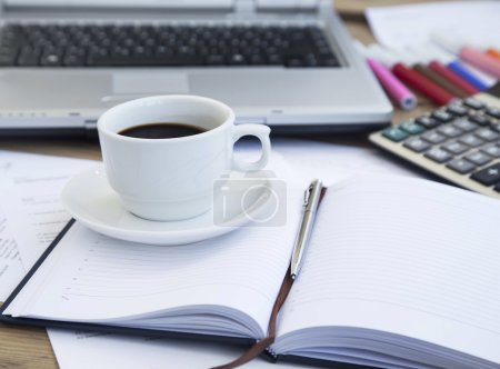 Foto de Taza de café en la oficina colocada sobre papeleo y agenda con pluma, calculadora y computadora portátil en el fondo - Imagen libre de derechos